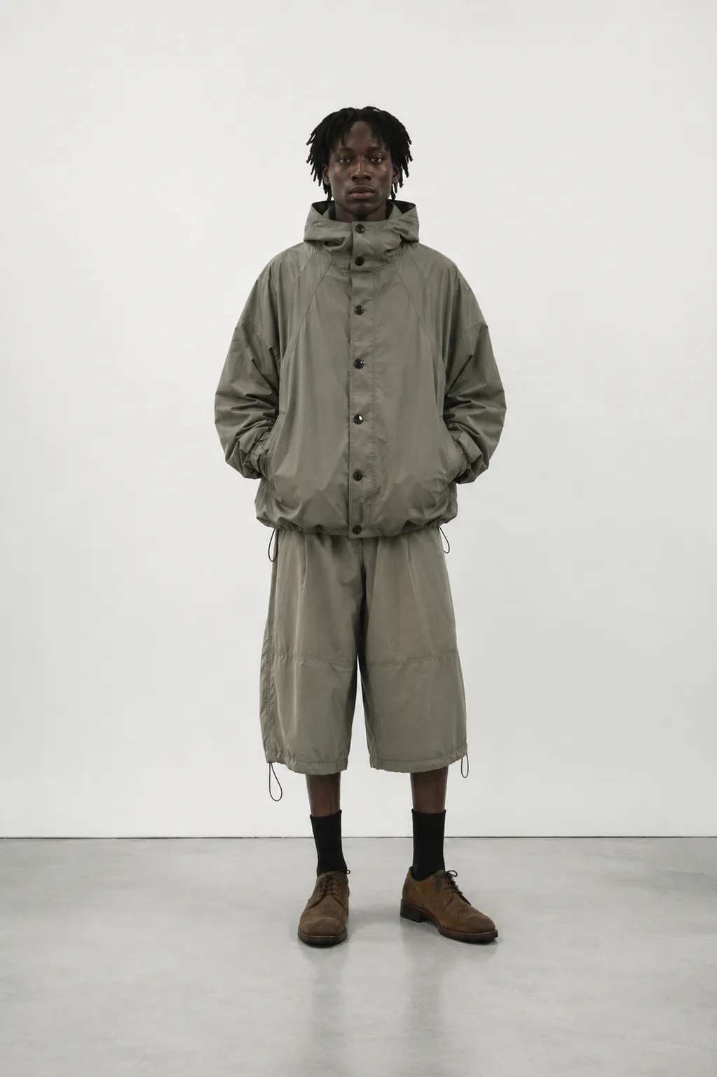 Mandlig model iført Orren Vale Mistral Hood Jacket i Sage Stone med matchende tekniske shorts, sorte strømper og brune lædersko.