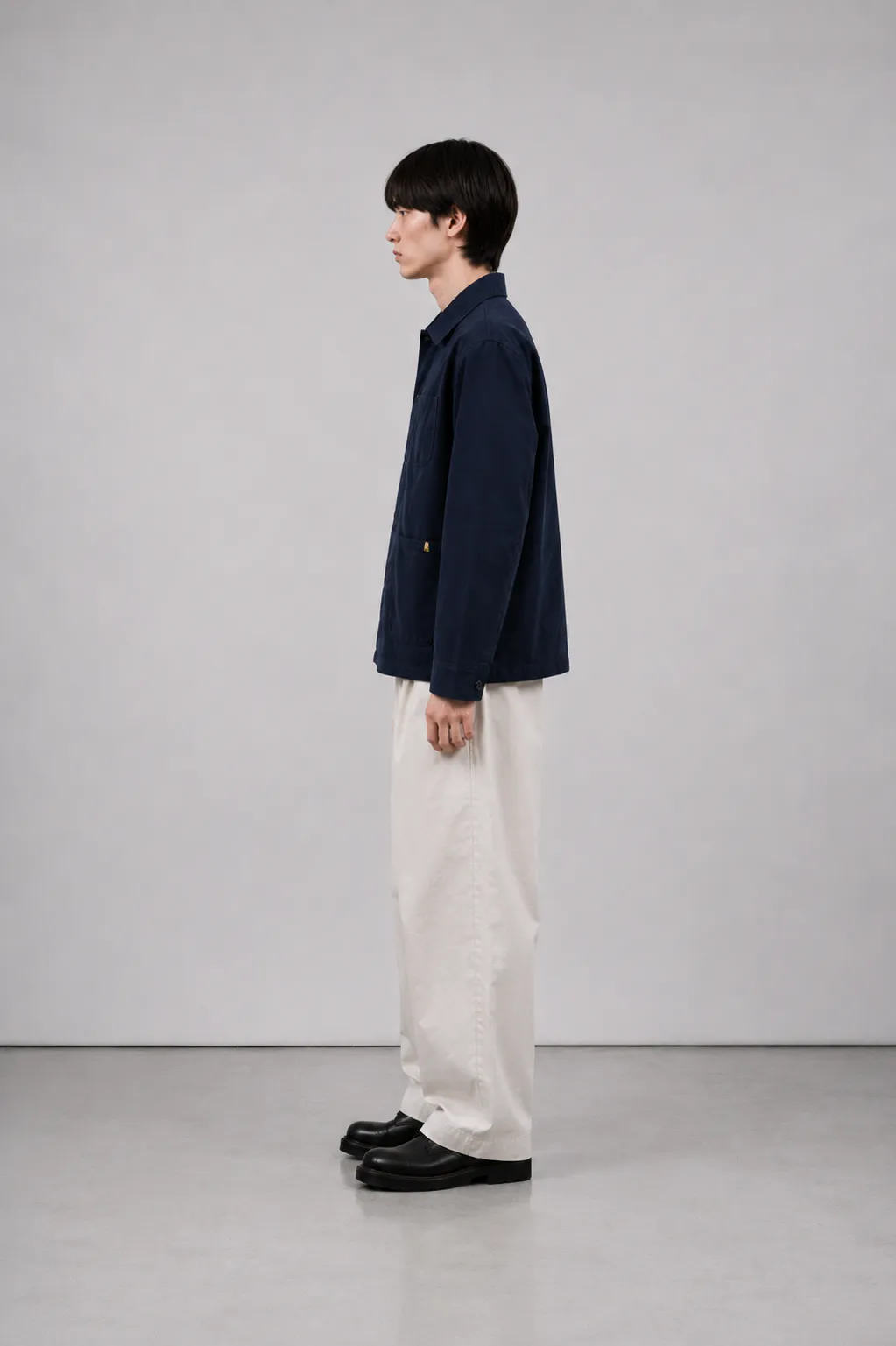 Mandlig model set fra siden iført Forvay Studio Marlo Work Jacket i Midnight Navy med hvid T-shirt, lyse bukser og sorte lædersko.