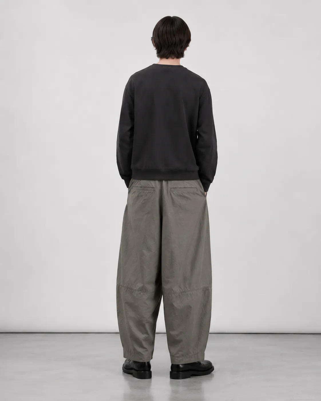 Mandlig model set bagfra iført Elvarn Studio Rivo Tech Pleat Trouser i Sage Stone med sort sweatshirt og sorte loafers.