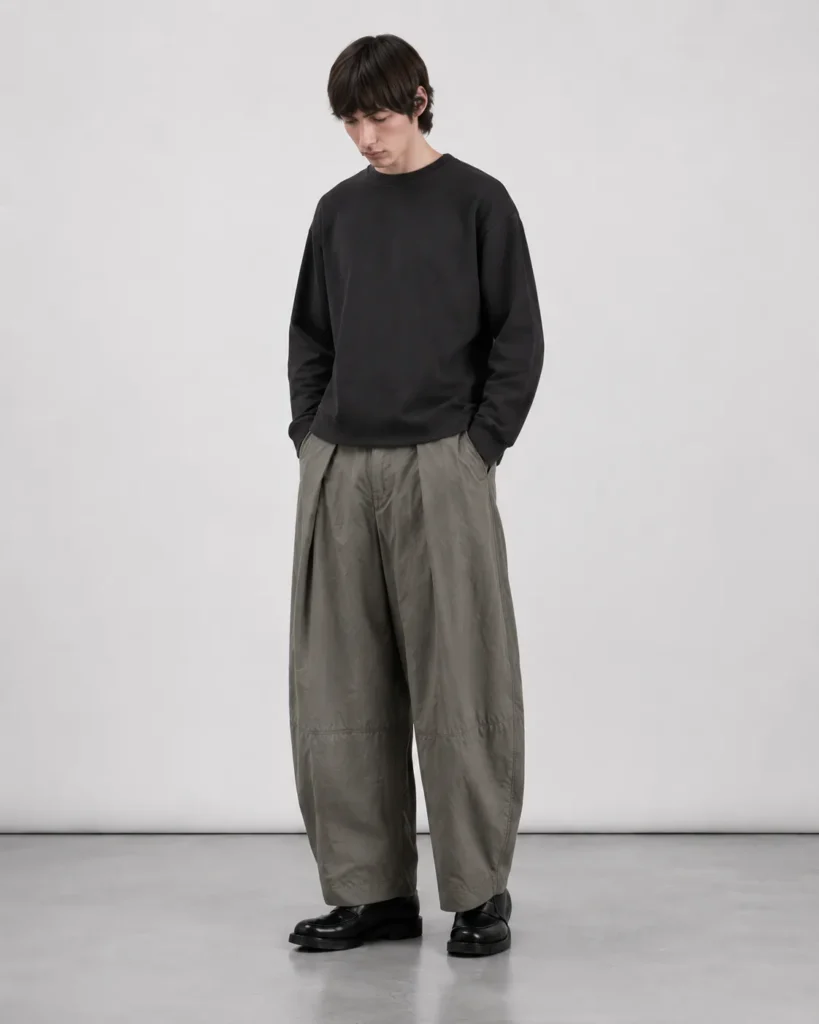 Mandlig model iført Elvarn Studio Rivo Tech Pleat Trouser i Sage Stone med sort sweatshirt og sorte loafers.