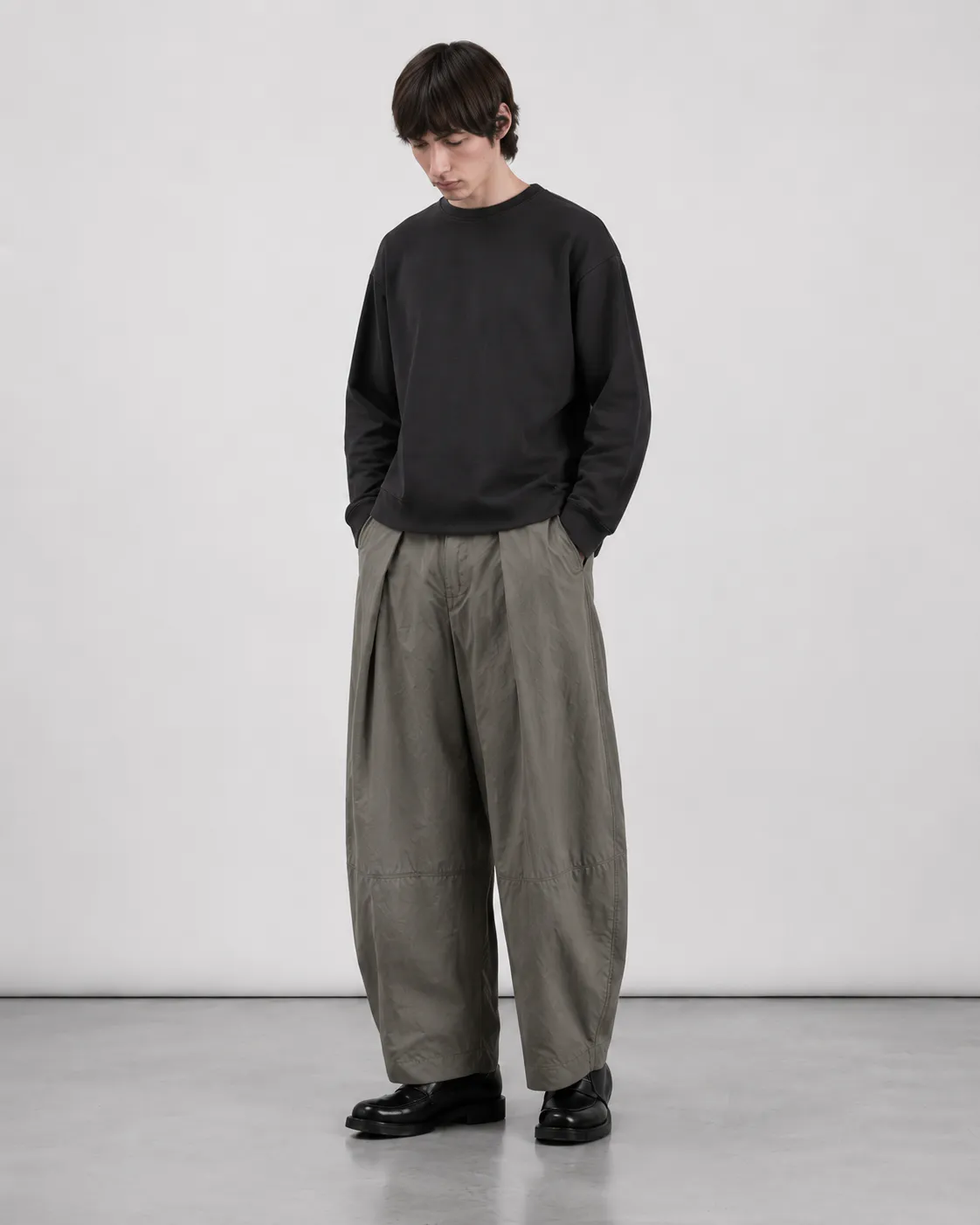 Mandlig model iført Elvarn Studio Rivo Tech Pleat Trouser i Sage Stone med sort sweatshirt og sorte loafers.