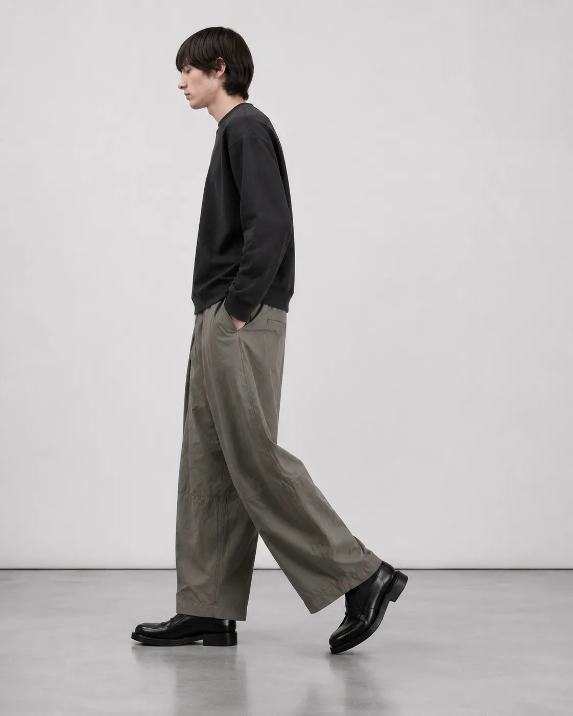 Mandlig model set fra siden iført Elvarn Studio Rivo Tech Pleat Trouser i Sage Stone med sort sweatshirt og sorte loafers.