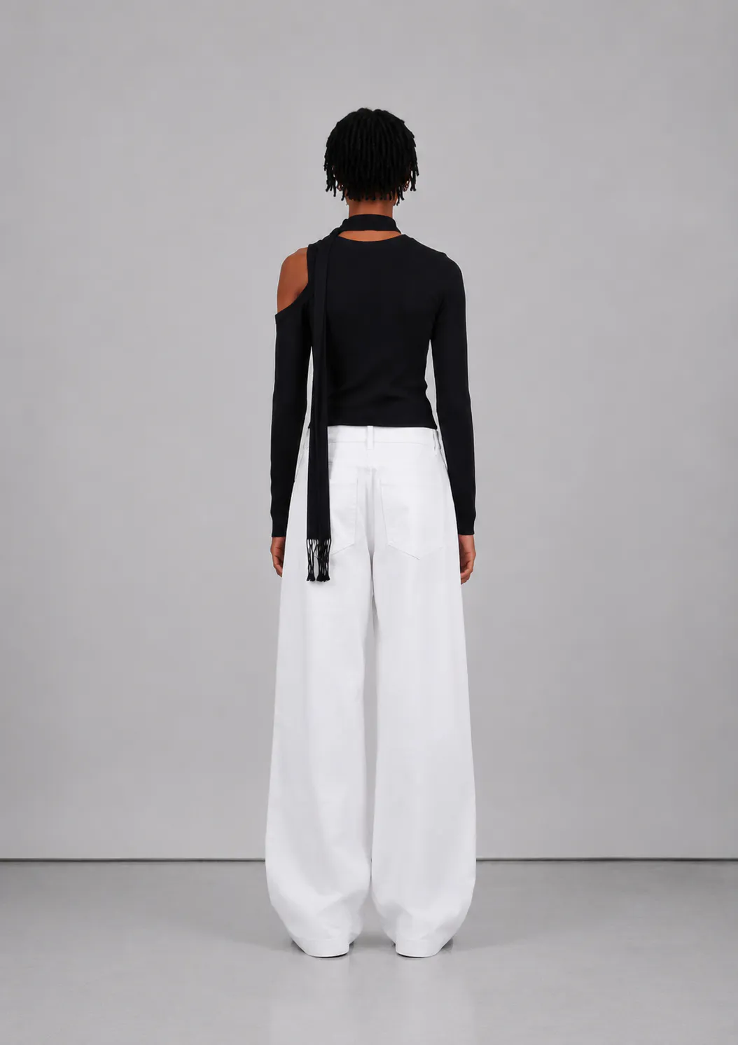 Kvindelig model set bagfra iført Veylan Studio Solenne Asym Top i Deep Black med hvide wide-leg bukser og sorte sandaler.
