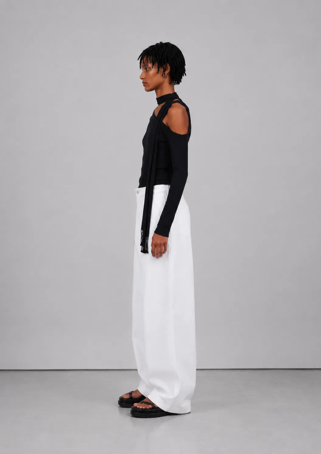 Kvindelig model set fra siden iført Veylan Studio Solenne Asym Top i Deep Black med hvide wide-leg bukser og sorte sandaler.